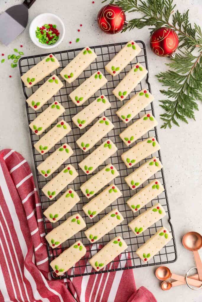 A cooling rack filled with 27 rectangular shortbread cookies, each decorated with green and red holly candies, and 6 docking marks in the middle.  Around the edges of the cooling rack are some cedar boughs, red festive Christmas balls, and candy sprinkles.