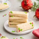 A stack of 5 rectangular short bread cookies on a small white dessert plate. Another shortbread cookie is laying on its side with it's top facing the camera. The two candy decorations of green holly and red berry can be seen. In the background are red festive Christmas balls and a teacup. A red linen is in the foreground.