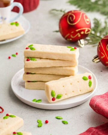 A stack of 5 rectangular short bread cookies on a small white dessert plate. Another shortbread cookie is laying on its side with it's top facing the camera. The two candy decorations of green holly and red berry can be seen. In the background are red festive Christmas balls and a teacup. A red linen is in the foreground.