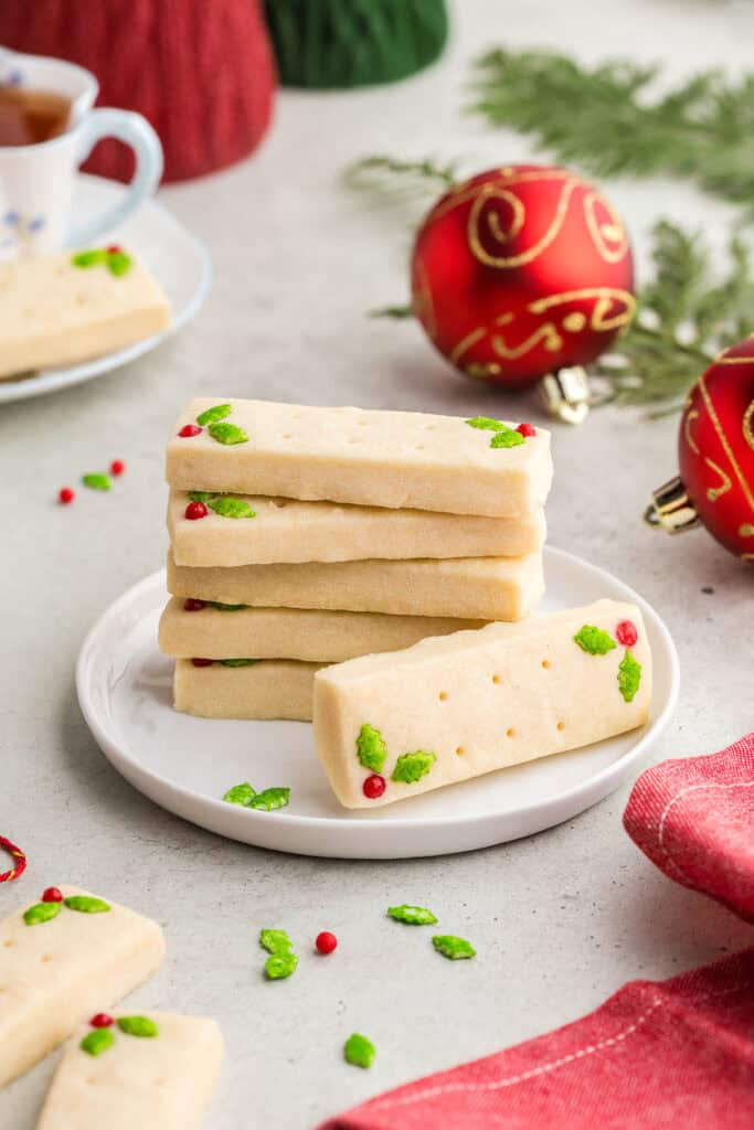 A stack of 5 rectangular short bread cookies on a small white dessert plate. Another shortbread cookie is laying on its side with it's top facing the camera. The two candy decorations of green holly and red berry can be seen. In the background are red festive Christmas balls and a teacup. A red linen is in the foreground.