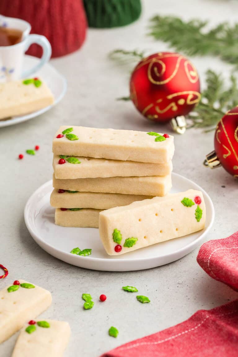A stack of 5 rectangular short bread cookies on a small white dessert plate. Another shortbread cookie is laying on its side with it's top facing the camera. The two candy decorations of green holly and red berry can be seen. In the background are red festive Christmas balls and a teacup. A red linen is in the foreground.