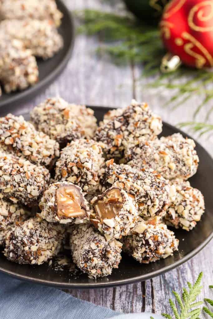 Side shot of a small black plate filled with pieces of almond roca. One piece at the front has been broken in two to show off the brown toffee inside. Sliced almonds are apparent inside the toffee, and the chocolate coating can also be identified. Finely chopped almonds are the final coating of these almond rocas.