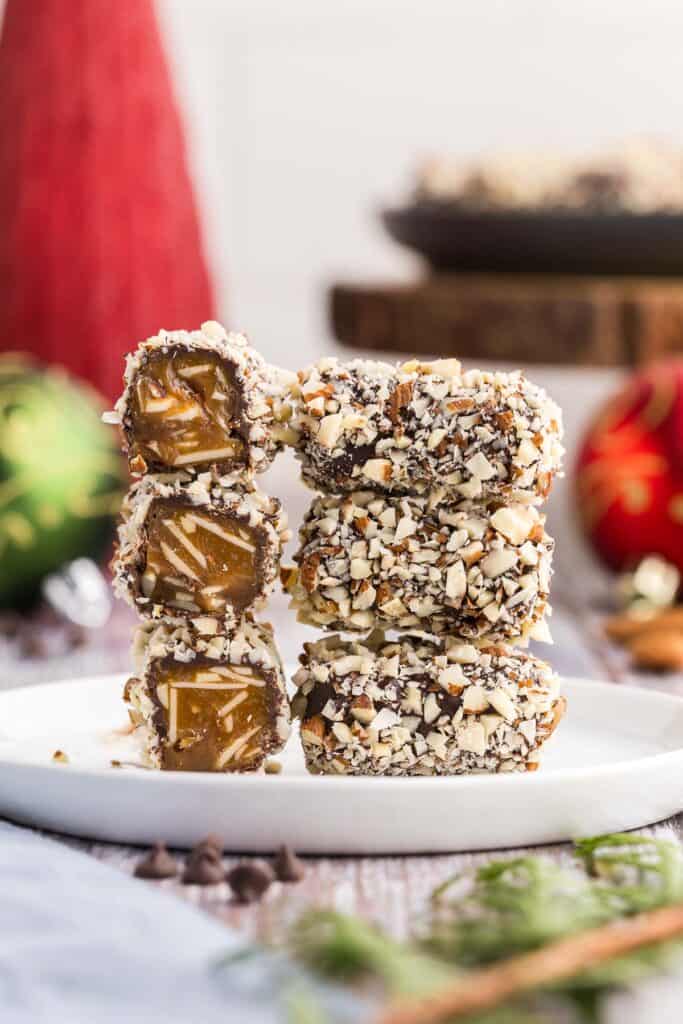 Side shot of two stacks of almond roca on a small white dessert plate.  The stack on the left is facing the camera with it's short ends.  The ends have been cut off so the interior is visible, a dark toffee with lots of sliced almonds are in the middle, then there is a thin coating of dark chocolate and finally a coating of chopped almonds on the outside.  The stack on the right is of three almond rocas stacked with their long sides facing the camera.  This shows the length of the almond roca to be about 1.5" and covered in chocolate and finally chopped almonds.