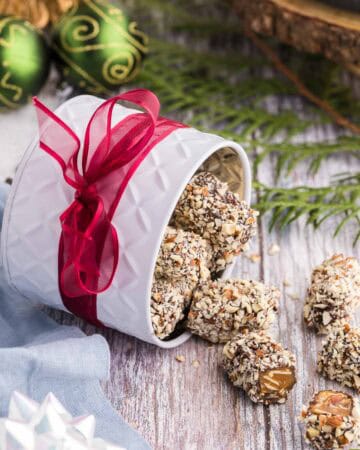 Side shot of a white tin with a red ribbon wrapped around it, laying on it's side and almond roca pouring out. One piece has been cut open revealing the golden brown toffee inside. Christmas bobbles are in the background and a white bow is in the foreground along with cedar boughs.