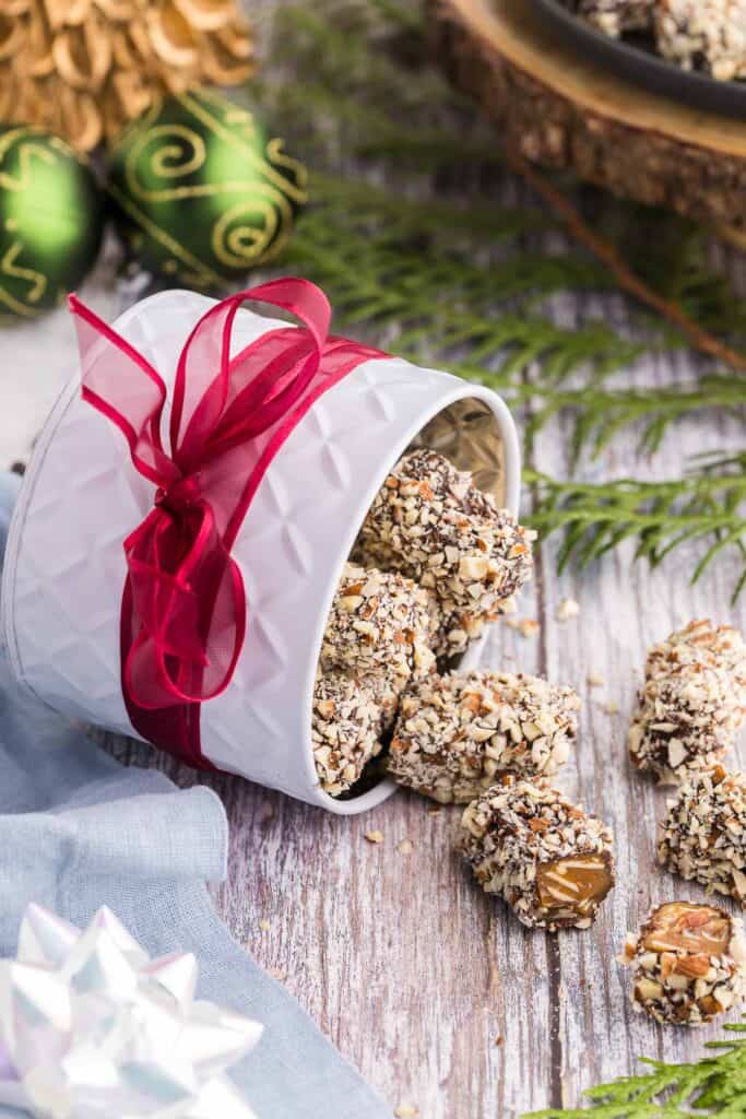 Side shot of a white tin with a red ribbon wrapped around it, laying on it's side and almond roca pouring out. One piece has been cut open revealing the golden brown toffee inside. Christmas bobbles are in the background and a white bow is in the foreground along with cedar boughs.