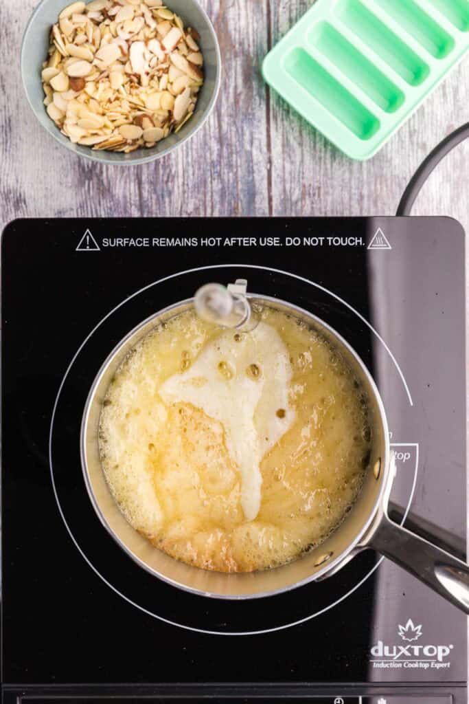 Overhead shot of a stainless steel saucepan with the light yellow colored sugar-butter mixture boiling away on top of an induction cooktop. The first step in making the toffee for almond roca.