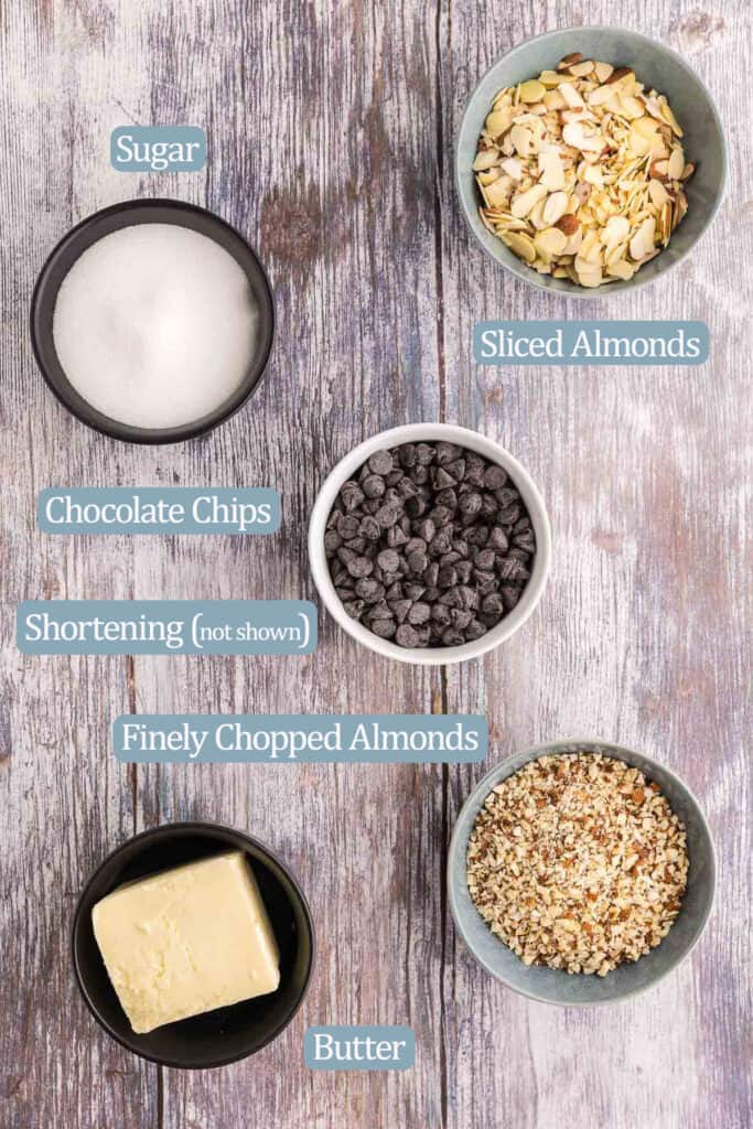 Overhead shot of the ingredients used in making Almond Roca. Ingredients are: white sugar, sliced almonds, chocolate chips, shortening, butter, finely chopped almonds.