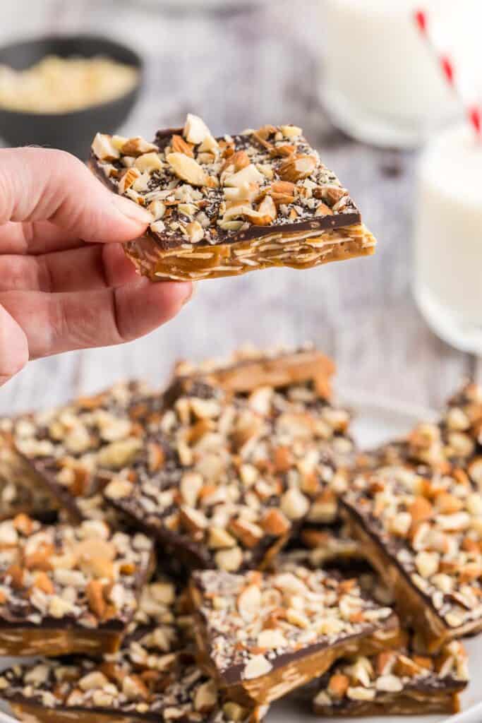Close up side shot of a piece of buttercrunch being lifted off a serving plate and held closer to the camera. The bottom toffee layer has sliced almonds dispersed throughout and the top layer is dark chocolate covered in chopped almonds.