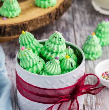 Side shot of a white circular gift tin wrapped around it's middle with a red ribbon. The lid is off and the tin is filled with green Christmas tree meringues. More meringue cookies are in the background on a wooden serving plater.