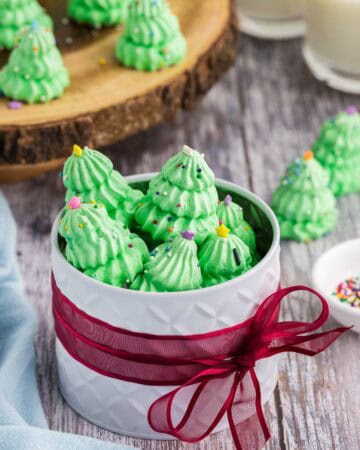 Side shot of a white circular gift tin wrapped around it's middle with a red ribbon. The lid is off and the tin is filled with green Christmas tree meringues. More meringue cookies are in the background on a wooden serving plater.