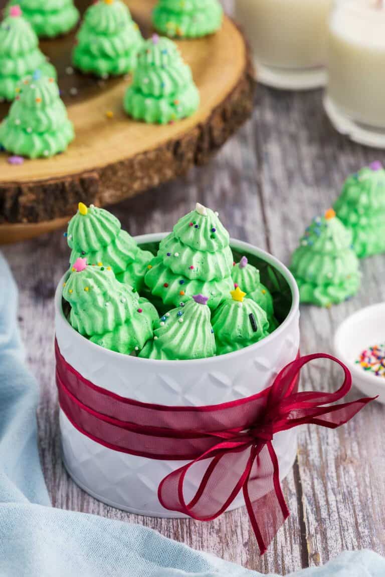 Side shot of a white circular gift tin wrapped around it's middle with a red ribbon. The lid is off and the tin is filled with green Christmas tree meringues. More meringue cookies are in the background on a wooden serving plater.