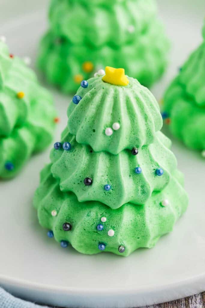 Close up side shot of a green Christmas Tree Meringue.  There are three layers to the tree.  Each layer is piped with a star tip giving each layer piped edges all the way around.  Each layer gets smaller as you go up, and the top layer is adorned with a yellow star sprinkle.  Blue and white pareills sprinkles have been sprinkled lightly all around the tree.  In the background three other similar trees are somewhat visible.