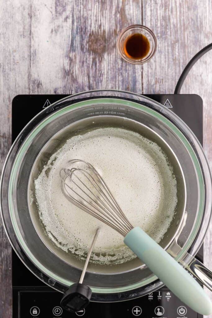 Overhead shot of a glass bowl on a double boiler with the egg whites, sugar, and cream of tartar being heated up to 170F  The mixture is slightly yellow and foamy.