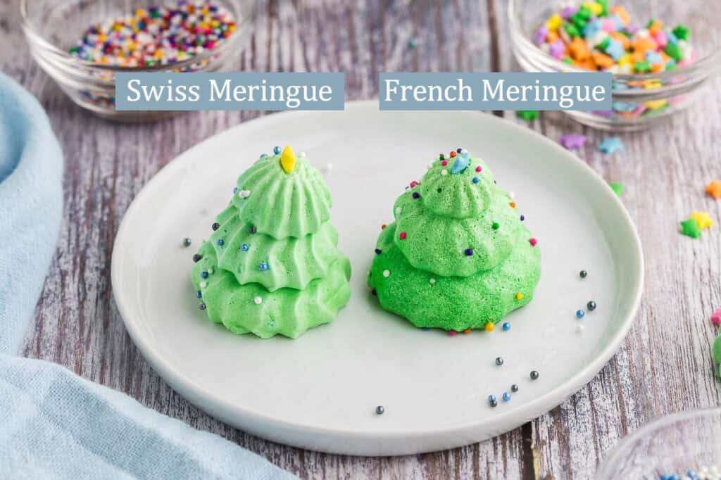 Shot of two Christmas Tree Meringues sitting side by side on a small white dessert plate.  The tree on the  right is made from Swiss Meringue and the piping ridges are clearly visible adding depth to the shape of the tree.  The tree on the right has been made from French meringue.  The French meringue tree is more smooth and ridges barely visible.  Pinch bowls of sprinkles are peeking in to the scene from the top corners.