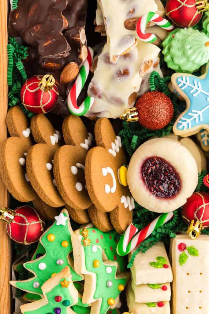 Close up overhead shot looking into a holiday cookie box. Cookies that can be seen are gingerbread men, green iced Christmas tree sugar cookies, shortbread, almond bark, and raspberry jam thumbprint cookies.