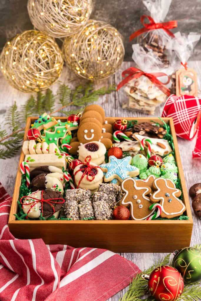 high angle shot looking down at a wooden box filled with Christmas cookies. Cookies include blue snowflake sugar cookies, chocolate peppermint cookies dipped in white chocolate, raspberry jam thumbprint cookies, gingerbread, almond roca to name a few. In the background are some cellophane bags of almond bark tied with a red bow.