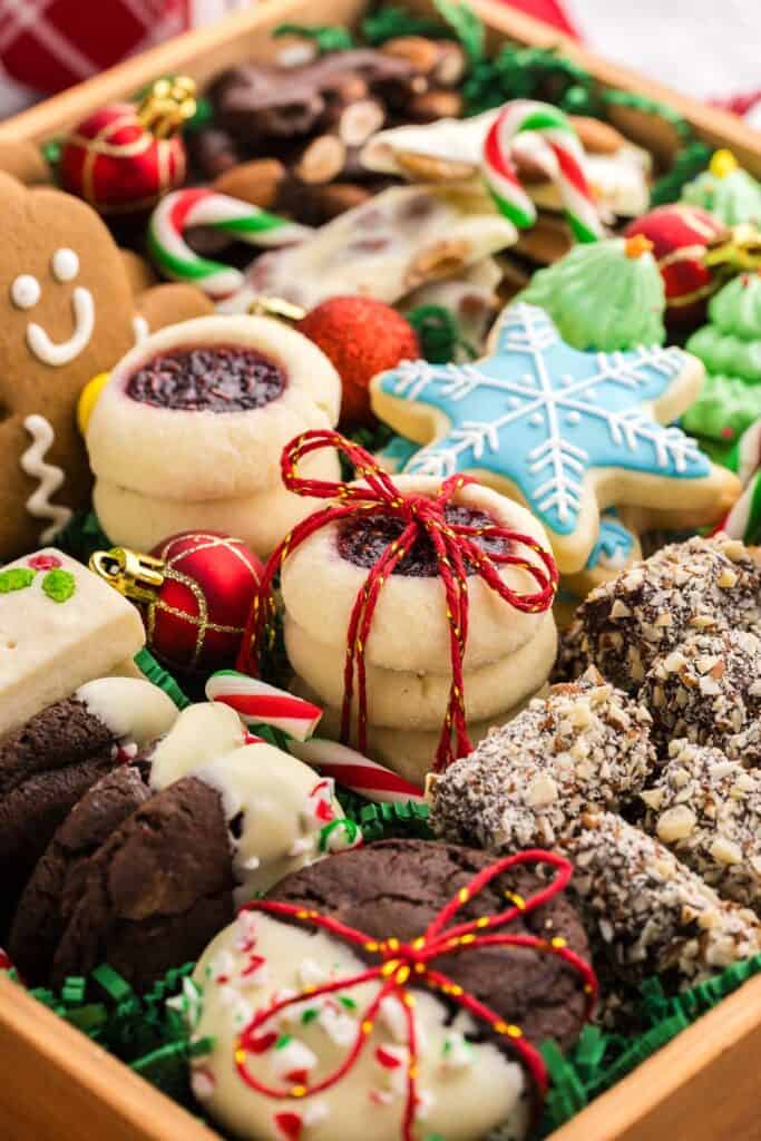 Close up side shot of a wooden box filled with many different types of Christmas Cookies. Upfront are chocolate peppermint cookies dipped in white chocolate, almond roca, raspberry thumbprints, blue snowflake sugar cookies, gingerbread men, green Christmas tree meringues, dark and white chocolate almond bark.