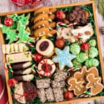 Overhead shot of a square wooden box filled with 8 different types of cookies. There are green Christmas tree sugar cookies, blue snowflake sugar cookies, gingerbread men and women, shortbread cookies, chocolate peppermint cookies, almond roca, raspberry thumbprint cookies tied with red string, white and dark chocolate almond bark, and green Christmas tree meringues.