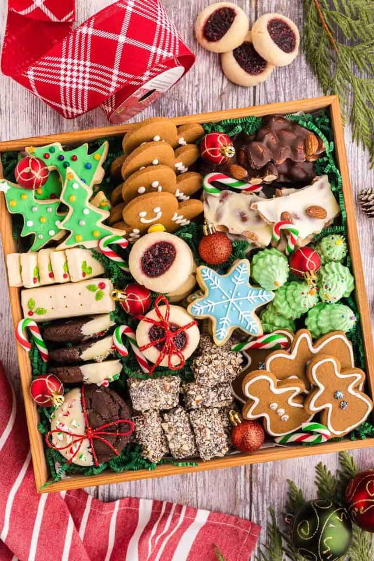 Overhead shot of a square wooden box filled with 8 different types of cookies. There are green Christmas tree sugar cookies, blue snowflake sugar cookies, gingerbread men and women, shortbread cookies, chocolate peppermint cookies, almond roca, raspberry thumbprint cookies tied with red string, white and dark chocolate almond bark, and green Christmas tree meringues.
