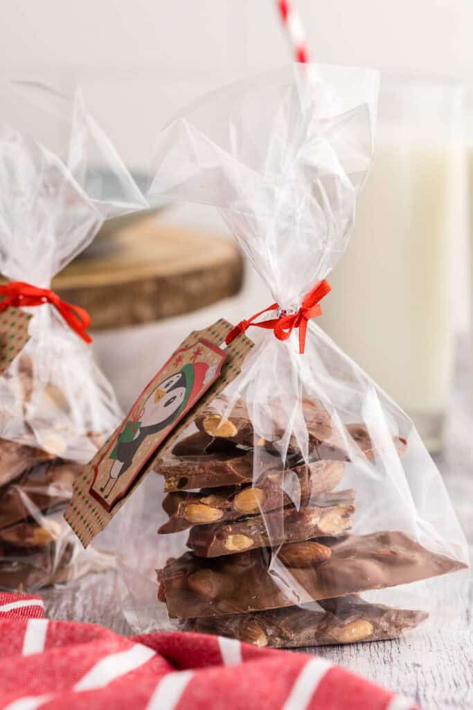 side shot of a cellophane gift bag filled with milk chocolate bark with almonds.  The bag is tied with a Christmas gift tag of a penguin.  In the background another gift bag is peeking in on the left, and on the right is a tall glass of milk with a red and white straw.