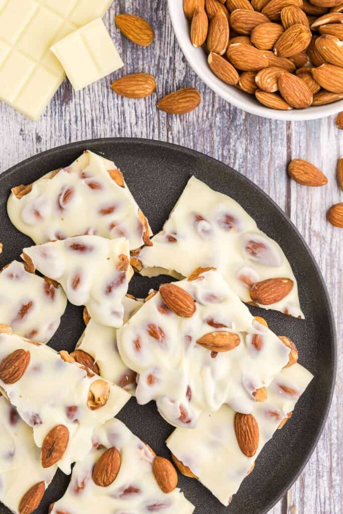 Overhead shot of a black ceramic plate filled with white chocolate almond bark.  The almond bark has been broken into large pieces, is a creamy off white color and studded with lots of almonds.  In the top left corner is a small bowl of almonds peeking in to the scene and there are almonds scattered around.