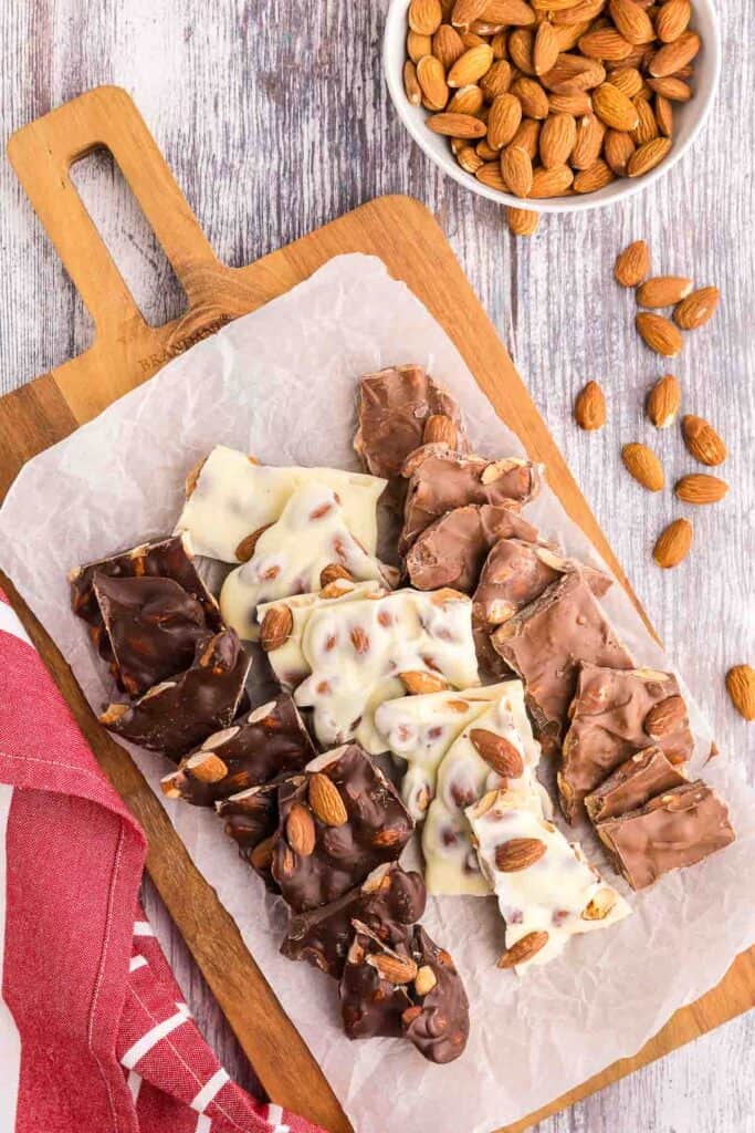 Overhead shot of a cutting board with white parchment paper.  On it are pieces of white almond bark, dark chocolate almond bark, and milk chocolate almond bark.  Each type is laid out in a line to form three lines of almond bark types.  In the upper corner of the picture is a small white bowl of almonds with some almonds spilled out.