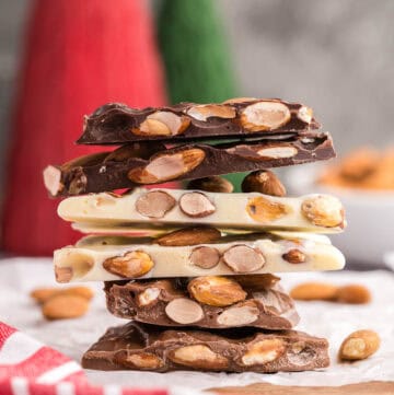 Side shot of a stack of almond bark. The bottom two pieces are milk chocolate almond bark, with a light brown, the middle two pieces are white chocolate almond bark with a creamy color, and the top two pieces are dark chocolate almond bark with a strong dark brown color. All types of bark are studded with lots of brown toasted almonds. This almond bark stack is sitting on white parchment paper on top of a wooden cutting board.