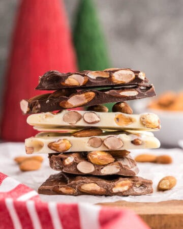 Side shot of a stack of almond bark. The bottom two pieces are milk chocolate almond bark, with a light brown, the middle two pieces are white chocolate almond bark with a creamy color, and the top two pieces are dark chocolate almond bark with a strong dark brown color. All types of bark are studded with lots of brown toasted almonds. This almond bark stack is sitting on white parchment paper on top of a wooden cutting board.