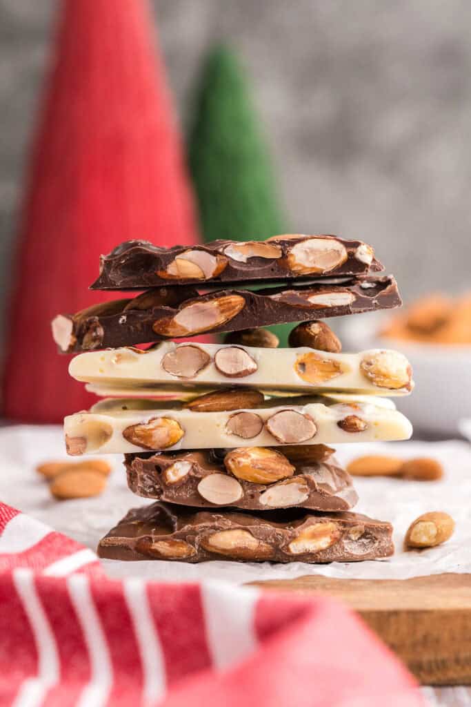 Side shot of a stack of almond bark. The bottom two pieces are milk chocolate almond bark, with a light brown, the middle two pieces are white chocolate almond bark with a creamy color, and the top two pieces are dark chocolate almond bark with a strong dark brown color. All types of bark are studded with lots of brown toasted almonds. This almond bark stack is sitting on white parchment paper on top of a wooden cutting board.