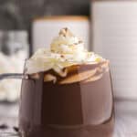 Side shot of a glass mug of dark chocolate hot chocolate. The mug is filled with dark chocolate hot cocoa and is topped with piped whipped cream and garnished with a dusting of cocoa powder. There are some chocolate chips scattered in the foreground.