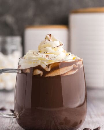 Side shot of a glass mug of dark chocolate hot chocolate. The mug is filled with dark chocolate hot cocoa and is topped with piped whipped cream and garnished with a dusting of cocoa powder. There are some chocolate chips scattered in the foreground.