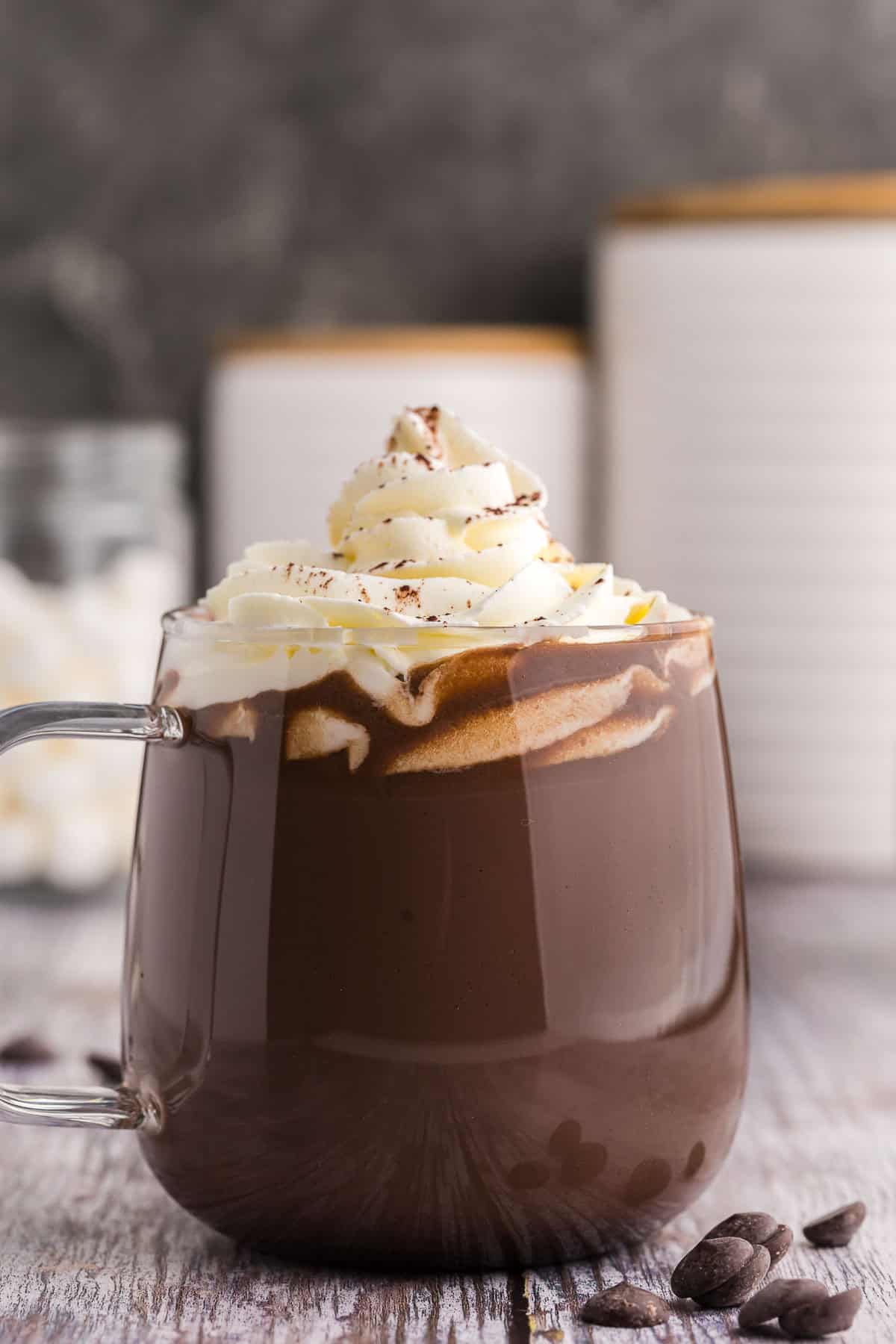 Side shot of a glass mug of dark chocolate hot chocolate. The mug is filled with dark chocolate hot cocoa and is topped with piped whipped cream and garnished with a dusting of cocoa powder. There are some chocolate chips scattered in the foreground.