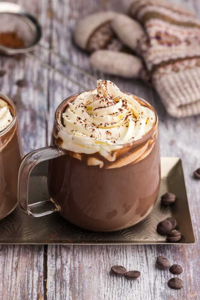 A glass mug of hot chocolate topped with lots of piped whipped cream and dusted with cocoa powder sits on a narrow rectangular serving tray. Another mug of the same hot cocoa is on the same serving tray but only a small portion of it is visible on the left of the main cup. In the background are some wool mittens and a sifter with some cocoa powder in it.