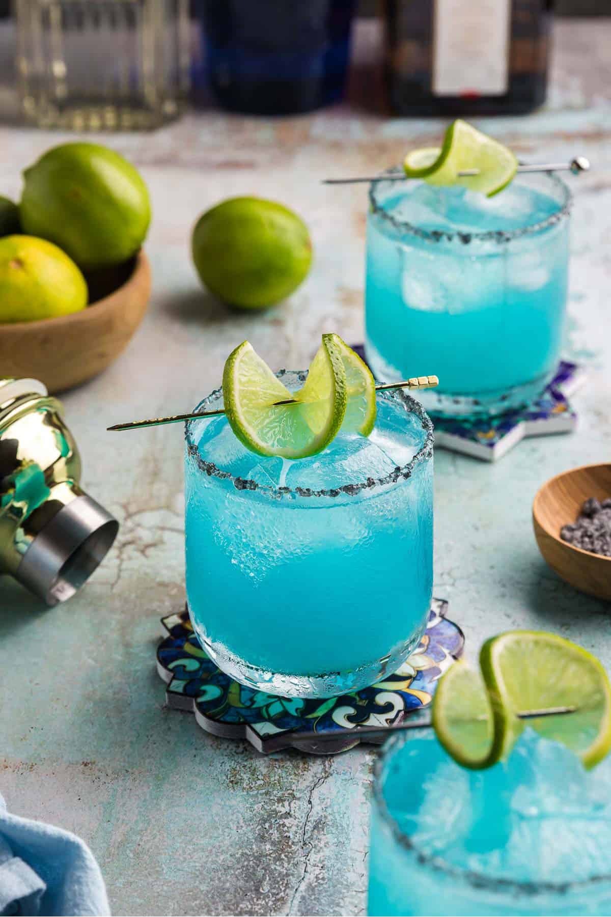 Three blue margaritas in low ball glasses.  Each one is rimmed with black salt and has a lime wheel corkscrew skewered on a cocktail pick.