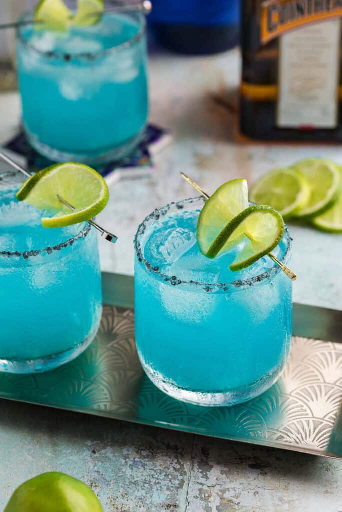 two blue margaritas in low ball glasses with black salt rims. A skewered lime wheel sits atop. A third blue margarita is in the background.