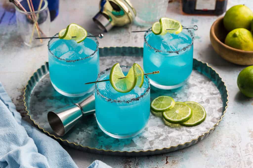 three blue margaritas on an oval metal serving tray. Each low ball glass is filled with ice and an ocean blue hue cocktail. The rims are covered lightly in black salt, and a lime wheel is skewered an laying on top of each glass.
