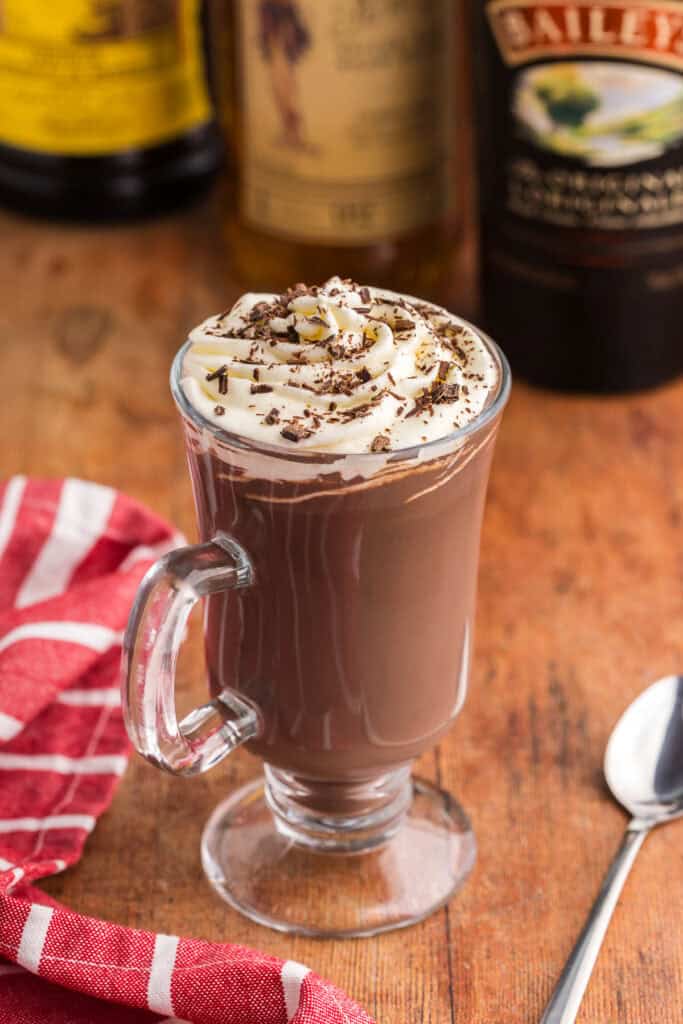 low angle shot of a pedestal-style glass mug filled with dark creamy spiked hot chocolate. The mug is topped with piped whipped cream and chocolate shavings. In the background are three bottles of Baileys, Kahlua, and spiced rum.