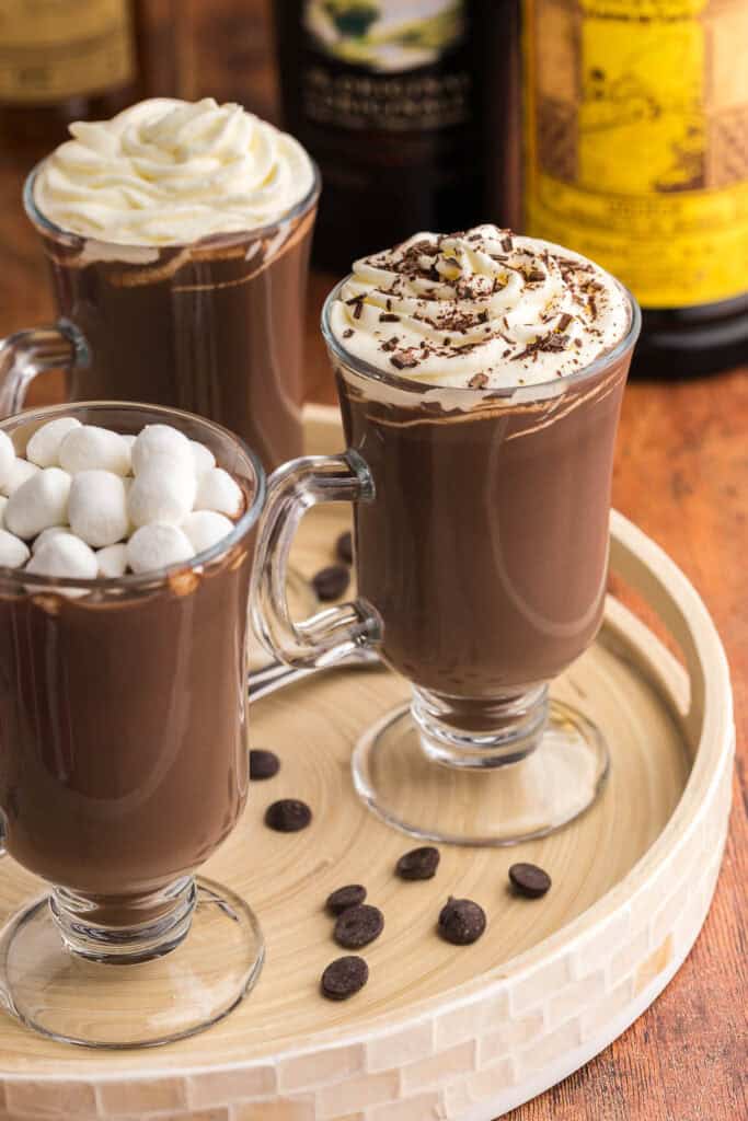 low angle shot of half a wooden serving tray with three glass mugs of spiked hot chocolate. Each one is topped differently. The one closest to the camera bottom left is topped with white mini marshmallows. The mug to the right and nearer the middle of the scene is topped with whipped cream and shaved chocolate. The third mug is towards the left and near the top of the photo is topped with just whipped cream.