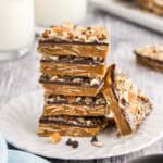 Side shot of a small plate with a stack of square buttercrunch pieces. Five pieces are in the stack and another piece is lying up against the stack. The brown toffee layer has lots of sliced almonds visible in it, and the chocolate layer can be seen on top with lots of chopped almonds attached to the top of the chocolate.
