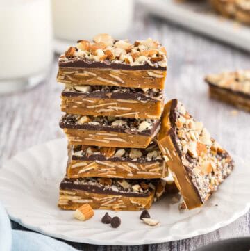 Side shot of a small plate with a stack of square buttercrunch pieces. Five pieces are in the stack and another piece is lying up against the stack. The brown toffee layer has lots of sliced almonds visible in it, and the chocolate layer can be seen on top with lots of chopped almonds attached to the top of the chocolate.