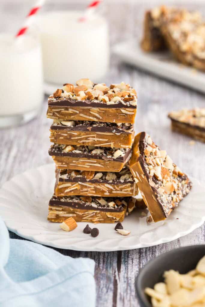 Side shot of a small plate with a stack of square buttercrunch pieces. Five pieces are in the stack and another piece is lying up against the stack. The brown toffee layer has lots of sliced almonds visible in it, and the chocolate layer can be seen on top with lots of chopped almonds attached to the top of the chocolate.