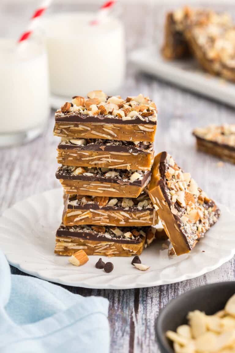 Side shot of a small plate with a stack of square buttercrunch pieces. Five pieces are in the stack and another piece is lying up against the stack. The brown toffee layer has lots of sliced almonds visible in it, and the chocolate layer can be seen on top with lots of chopped almonds attached to the top of the chocolate.