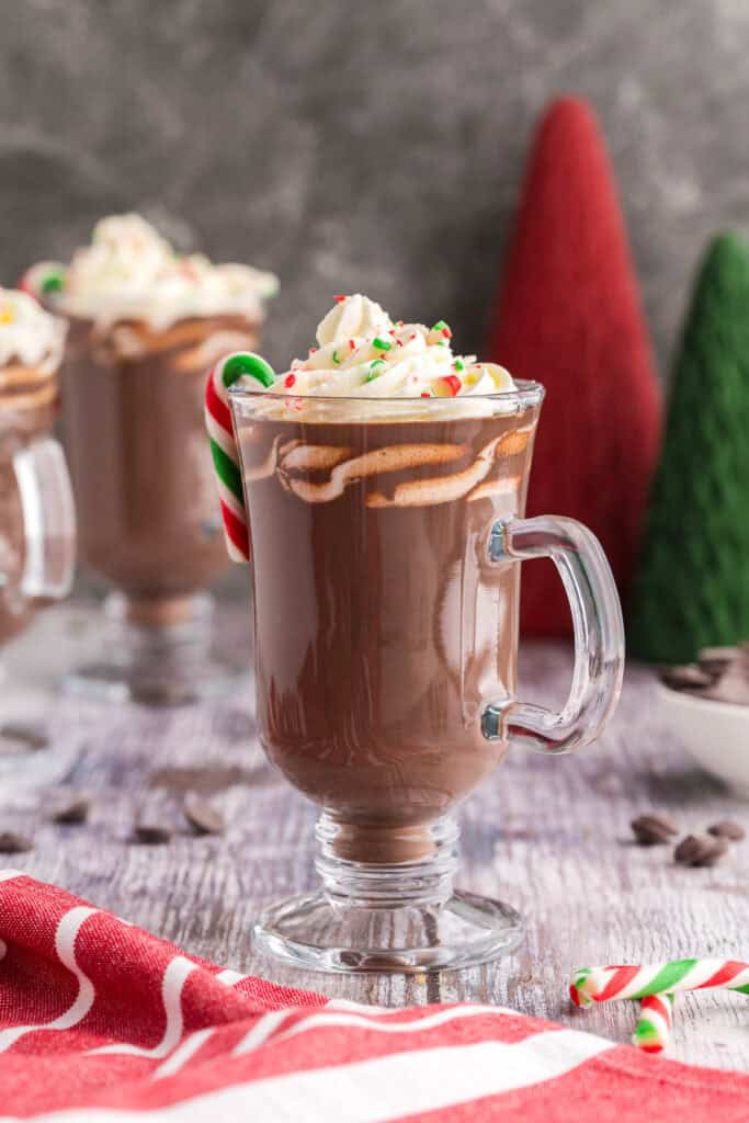 Side shot of a glass pedestal-style mug with dark brown peppermint hot chocolate.  Topped with whipped cream and crushed candy canes, and has a small candy cane hanging on the edge opposite the handle.  In the background are two more mugs, and a red and green Christmas tree shaped decoration.  Chocolate chips and small candy canes are sparingly scattered around.