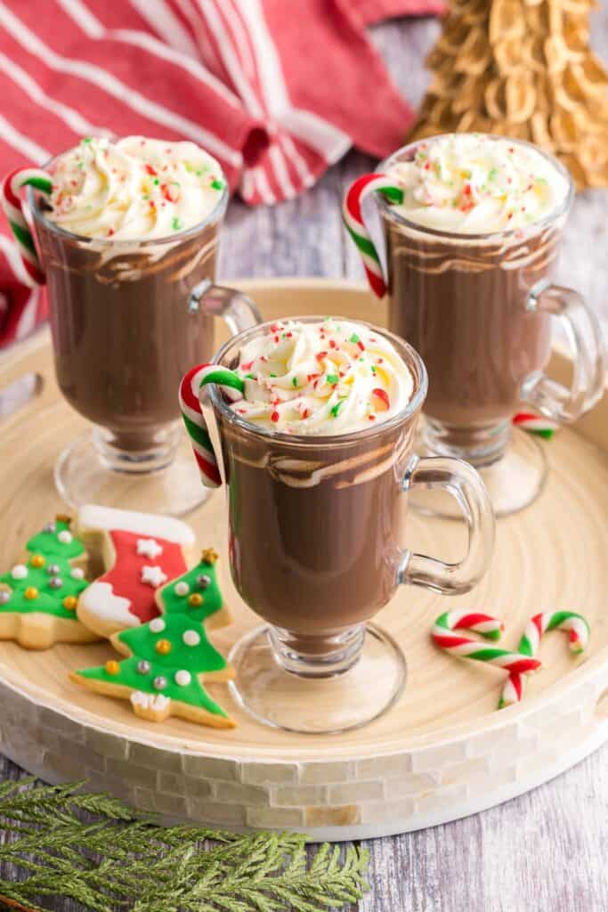 A beige round wooden serving tray has three glass mugs of peppermint hot chocolate.  Each one a dark brown with piped whipped cream and sprinkled with crushed candy canes.  Some Christmas sugar cookies and small candy canes are also on the tray.