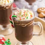 low oblique angle of a glass pedestal style mug filled with dark brown hot chocolate. On the top is creamy piped whipped cream and sprinkles of crushed cane. There is also a green, red, and white small candy cane hanging off the edge of the drink, and more little candy canes scattered on the serving tray. Two other peppermint hot chocolates are peeking into the scene on the left and the top.