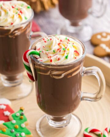 low oblique angle of a glass pedestal style mug filled with dark brown hot chocolate. On the top is creamy piped whipped cream and sprinkles of crushed cane. There is also a green, red, and white small candy cane hanging off the edge of the drink, and more little candy canes scattered on the serving tray. Two other peppermint hot chocolates are peeking into the scene on the left and the top.