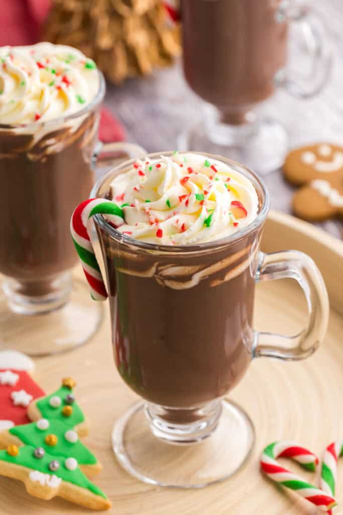 low oblique angle of a glass pedestal style mug filled with dark brown hot chocolate.  On the top is creamy piped whipped cream and sprinkles of crushed cane.  There is also a green, red, and white small candy cane hanging off the edge of the drink, and more little candy canes scattered on the serving tray.  Two other peppermint hot chocolates are peeking into the scene on the left and the top.