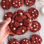 Overhead shot of a red velvet cake cookie being held up close to the camera. In the background is the cooling rack filled with more cookies. The cookie being held up has a crinkled surface and is studded with lots of white chocolate chips.