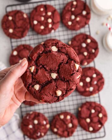 Overhead shot of a red velvet cake cookie being held up close to the camera. In the background is the cooling rack filled with more cookies. The cookie being held up has a crinkled surface and is studded with lots of white chocolate chips.