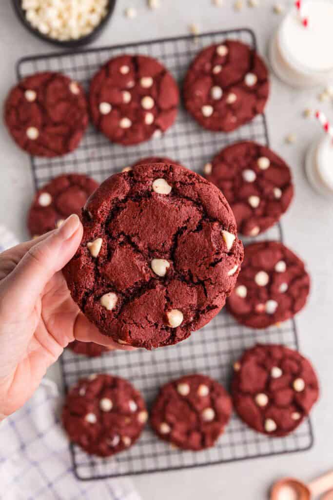 Overhead shot of a red velvet cake cookie being held up close to the camera. In the background is the cooling rack filled with more cookies. The cookie being held up has a crinkled surface and is studded with lots of white chocolate chips.